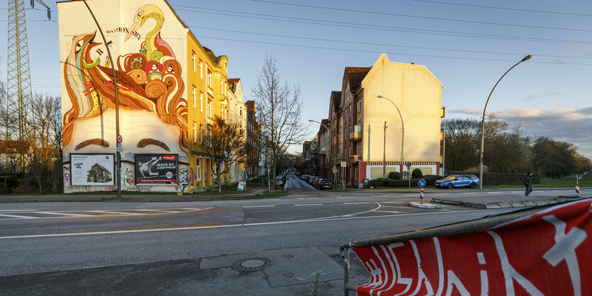 Die Sonne scheint auf ein Eckhaus in Hamburg-Wilhelmsburg, die Fassade ist bunt angemalt. Auf einem Banner steht "115 bleibt"