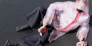 Ein Man sitzt nach hinten gebeugt auf dem Boden. Er trägt japanische Kleidung, sein Gesicht ist von einer Kapuze verdeckt