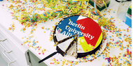 Ein angeschnittener Kuchen mit der Aufschrift "Berlin University Alliance" und Konfetti auf einem Schreibtisch