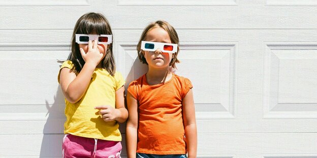 Zwei kleine Mädchen mit 3D Brille lehnen im Sonnenschein an der Wand