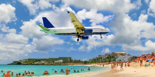 Flugzeug vor blauem Himmel mit Schäfchenwolken fliegt niedrig über Sanstrand mit Urlaubern und türkisblauem Meer