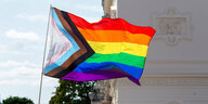 Eine Regenbogenflagge weht im Wind. Auch die Farben der trans* Flagge, weiß,rosa und blau, sind in einem Dreieck auf der linken Seite der Flagge zu sehen.