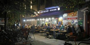 Eine große Gruppe von Menschen sitzen vor einem Kiosk auf Bänken.