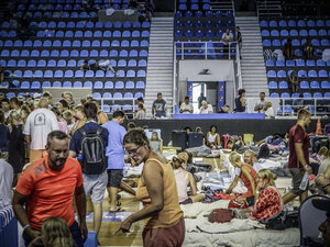 Menschen lagern auf dem Boden eines Stadions, telefonieren, gehen zum Eingang, schauen sich hilfesuchend um