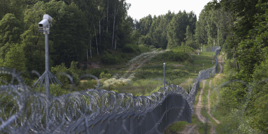 Ein Stacheldrahtzaun durchfurcht einen Wald, darüber Überwachungskameras