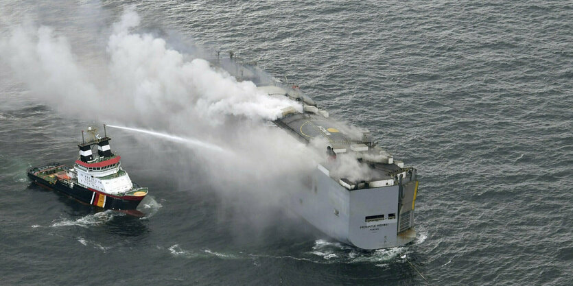 eine Luftaufnahme von einem rauchenden Schiff auf dem Meer, links daneben ein Löschschiff