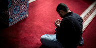 Ein Mann sitzt im Schneidersitz auf einem roten Teppich in einer Moschee. Er hält seine Hände in einer Gebetshaltung vor seinen Oberkörper.