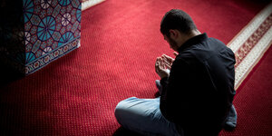 Ein Mann sitzt im Schneidersitz auf einem roten Teppich in einer Moschee. Er hält seine Hände in einer Gebetshaltung vor seinen Oberkörper.
