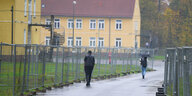 Zwei Menschen laufen zwischen Zäunen eine Straße entlang, im Hintergrund stehen gelbe Gebäude