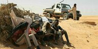 Vier Männer sitzen in der Wüste vor einem staubigen Buschwerk, hinter ihnen steht ein libyscher Soldat vor seinem Geländewagen