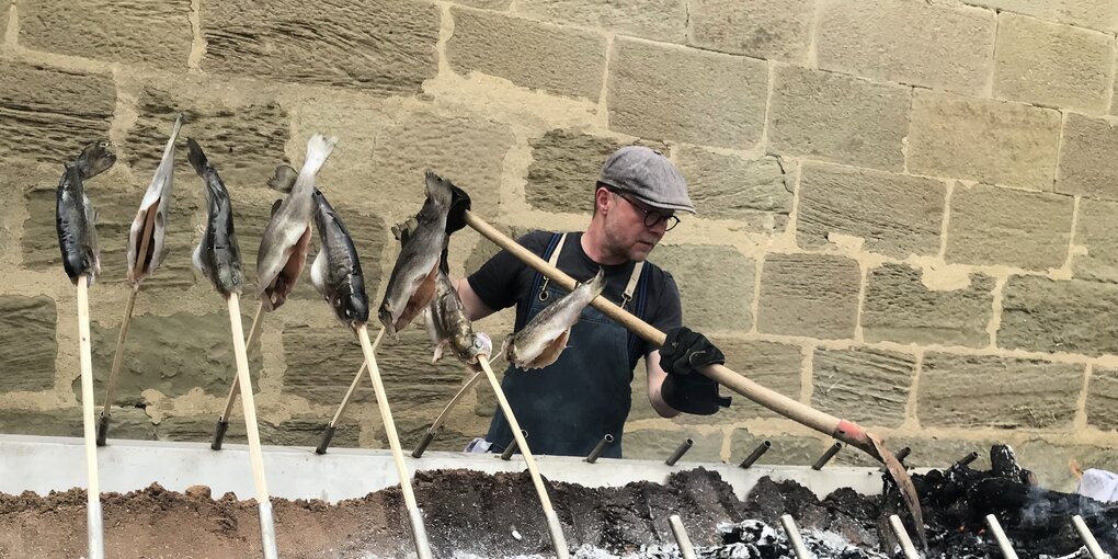 Ein Mann steht an einem riesigen Grill, über dem Fische brutzeln