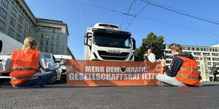 Klimaaktivist:innen der Letzten Generation sitzen in Warnwesten und mit einem Transparent auf einer Straße und hindern einen Lkw am Durchfahren.