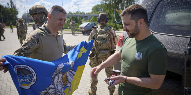 Ukrainischer Präsident Selenski mit Soldaten, die ihm eine blau-gelbe Flagge zeigen.