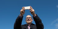 Thorsten Albig hält vor blauem Himmel ein Smartphone mit beiden Händen hoch