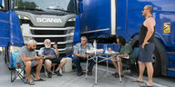 Männer sitzen auf Stühlen vor einem blauen Lkw. Ein Mann rechts steht.Auf einem Campingtisch liegt Essen und Trinken