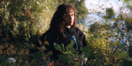 Eine junge Frau steht in der Natur etwas verdeckt von Büschen im Vordergrund