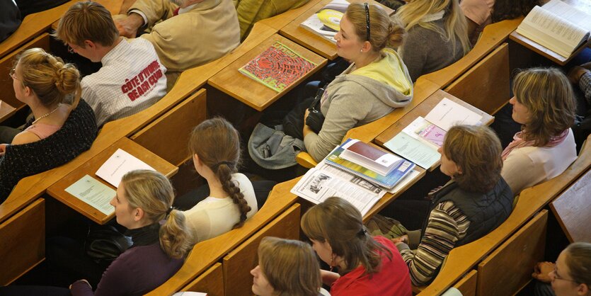 Studierende sitzen in einem Hörsaal. Aufgeschlagene Bücher liegen auf den Tischen.