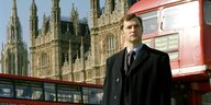 Ein Mann im Mantel mit Anzug und Krawatte steht in London vor einem Bus, im Hintergrund Westminster Abbey
