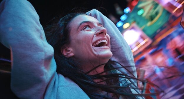 Eine junge Frau mit langen dunklen Haaren lacht und wirft die Hände in die Luft vor einer unscharfen "Rummel" Kulisse