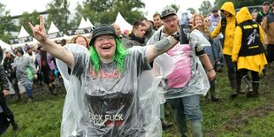 Eine Frau mit Regenschutz, grünen Haaren geht gut gelaunt mit anderen Festivalbesuchern über die matschige Wiese