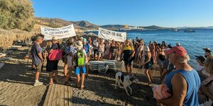 Griechische Protestplakate an einem Strand, wirres Durcheinander zwischen gestapelten Liegen im Sand