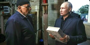 Wladimir Putin hält ein dickes Buch in der Hand und spricht mit einem älteren Mann, der eine schwarze Mütze trägt. Beide stehen in einem eher schmucklosen Raum der Moschee