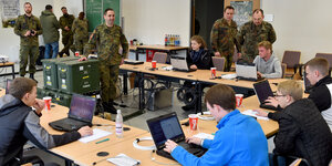 In einem Klassenraum stehen Soldaten in Bundeswehruniformen. An Tischen sitzen Jugendliche, die auf Laptops tippen.