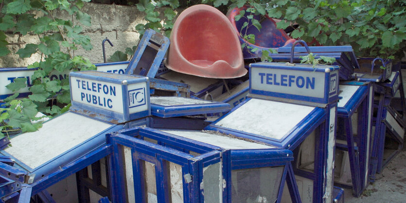 Aufnahme einer Reihe von ausrangierten Telefonhäusschen, die im Freien übereinander gestapelt gelagert sind. Die Häusschen haben blaue Metallstrukturen und silberne Wände, auf einigen ist das Wort "Telefon" zu lesen, auf anderen "Telefon Public".