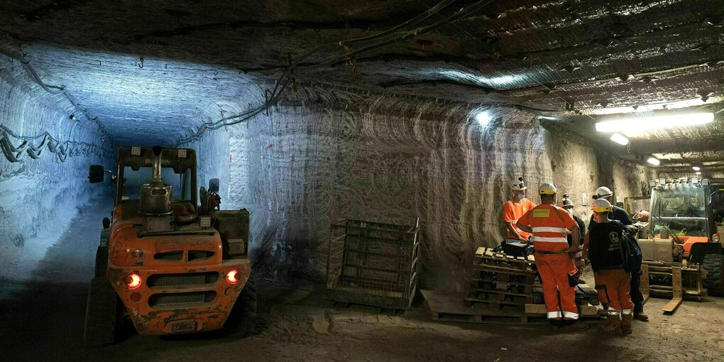 Blick in den Gang eines Stollens in dem ein orangefarbenes Fahrzeug steht, die Männer im Nebenstollen tragen leuchtende Schutzkleidung und Helme