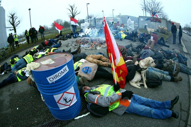 Protest vor den Toren von Stocamine, die Demonstranten liegen auf dem Boden und blockieren damit die Einfahrt. Im Vordergrund steht ein blaues Fass auf dem das Wort Chrome steht