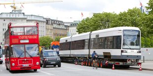 Eine weiße Straßenbahn wird auf einem LKW-Anhänger transportiert, daneben fährt ein roter Bus