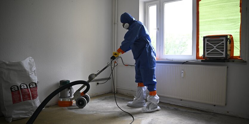 Ein Mann im Schutzanzug benutzt einen großen Staubsauger in einem leeren Raum mit Fenster