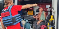 Ein älterer Mann sitzt in einem Rettungsfahrzeug. Ein Rot-Kreuz-Helfer steht draußen und zeigt mit dem Finger in Richtung des Fahrzeuginnenraums