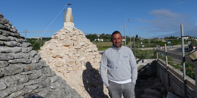 Ein junger Mann steht in der Abendsonne vor einem Trulli, sein Schatten wird auf das rundliche Bauwerk geworfen