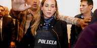 Ein Mann schirmt Andrea Gonzáles von hinten ab, sie trägt eine schussischere Weste und blickt nach unten.