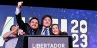 Javier Milei steht zusammen mit einem anderen Mann und einer Frau an einem Redepult und sie lächeln triumphierend in die Kamera.