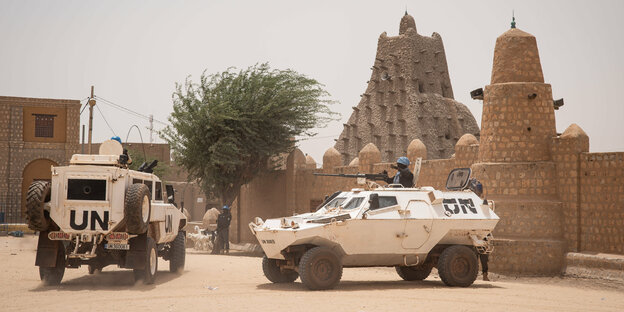 Zwei Militärfahrzeuge mit UN-Aufschrift stehen vor Steingebäuden in sehr sandiger Umgebung