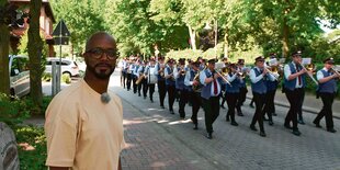 Yared Dibaba steht auf einer Straße auf der eine Blaskapelle vorbei geht.