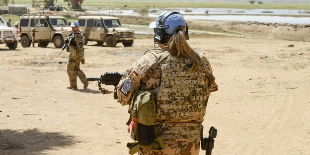Soldatin mit Pferdeschwanz und Camouflage, sandiger Untergrund, Jeeps im Hintergrund