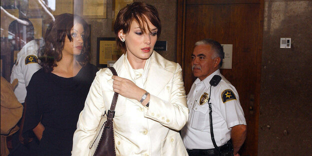 Winona Ryder im weißen Jacket legt sich eine Handtasche über die Schulter, Uniformierte im Hintergrund