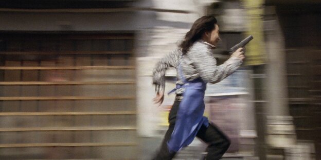 Ein Mann, der eine blaue Schürze trägt, rennt in Richtung der rechten Bildhälfte. Er hält einen Pistole in der Hand.