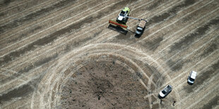 Ein Feld aus der Vogelperspektive. Man sieht Menschen, eine Maschine und Fahrzeuge. Unten ein kraterähnliches Loch.