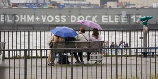 Passanten schützen sich mit Regenschirmen auf einer Sitzbank an den Hamburger Landungsbrücken vor dem Regen.