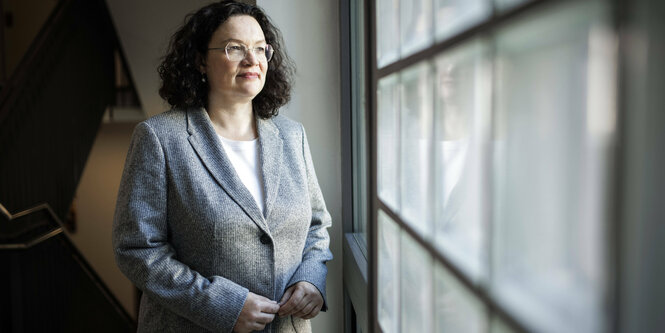 Andrea Nahles in einer Art Blazer aus Wolle, nachdenklich aus einem gekachelten Fenster schauend