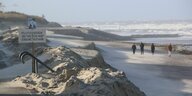 Spaziergänger gehen im Sturm auf der Insel Wangerooge am Strand entlang, im Vordergrund ist ein Schild zu sehen, auf dem steht: Flutschäden, betreten auf eigene Gefahr.