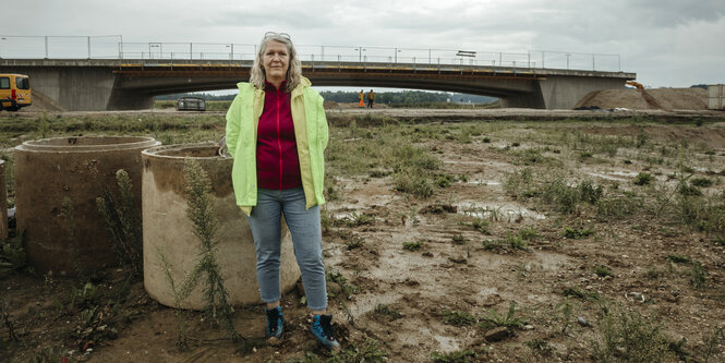 Eine Frau in gelber Regenjacke steht im Matsch, dahinter eine Brücke