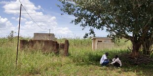 Zwei Männer sitzen neben einer Hausruine im SChatten eins Baumes und reden miteinander