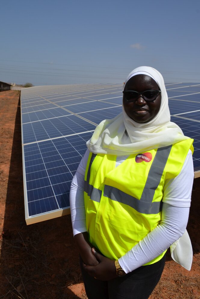 Person mit Gelbweste, Kopftuch und Sonnenbrille vor einer Solaranlage.