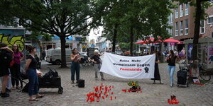 Auf einem städtischen, belebten Platz stehen Aktivistinnen mit Kerzen und einem Transparent, Aufschrift: Keine Mehr. Gemeinsam gegen Feminizide.