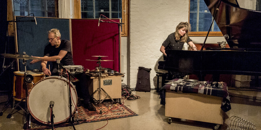 Burkhard Beins und Anaïs Tuerlinckx bei einem Konzert im Morphine Raum in Berlin. Beins sitzt links im Bild am Schlagzeug. Tuerlinckx spielt einen Flügel im Stehen. Im Hintergrung sind Fenster zu sehen, durch die der Abendhimmel scheint.